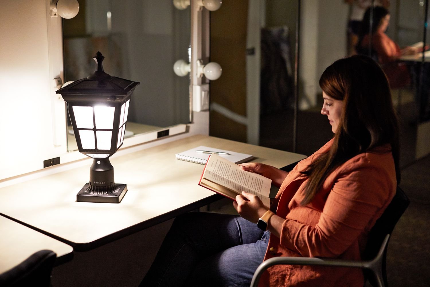 Woman sitting in a chair reading a book next to a table displaying a Kemeco Solar Post Light with Mount